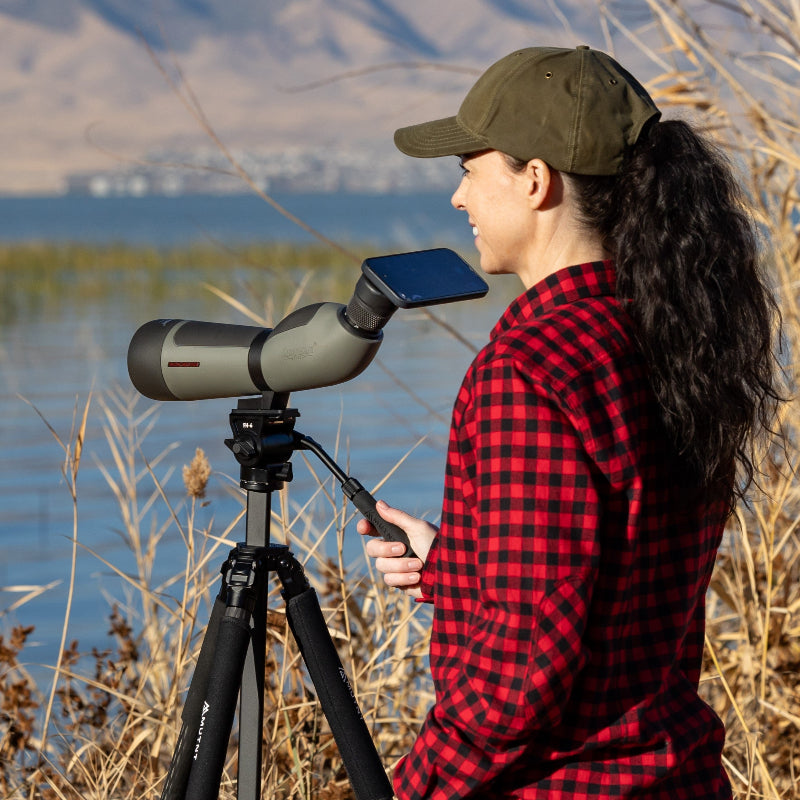 Person using a spotting scope by a body of water with mountains in the background